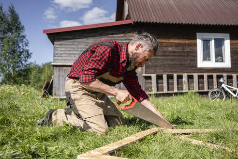 Carpenter working with hand saw cutting plank for playhouse on sunny day - VPIF02832