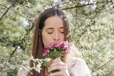 Frau riecht an frischen rosa Blumen, während sie im Frühling in einem Park unter Bäumen steht - RDGF00072