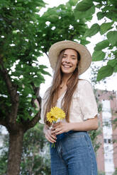 Smiling young woman wearing hat and holding yellow flowers while standing in park - RDGF00077