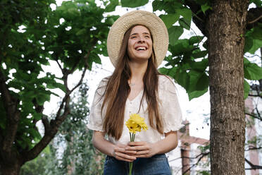 Lächelnde Frau schaut weg, während sie gelbe Blumen in einem öffentlichen Park im Frühling hält - RDGF00078