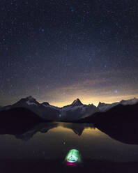 Starry night sky over illuminated tent pitched on shore of Bachalpsee lake at night - MALF00144