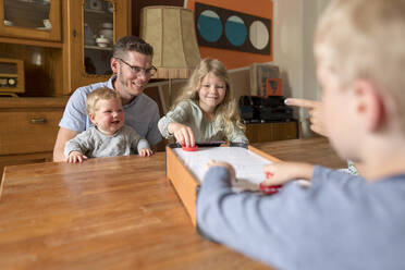 Smiling family watching kids playing air hockey on dining table at home - MOEF03082