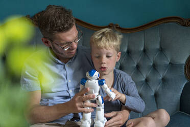 Cute blond boy looking at robot held by father while sitting on sofa in living room - MOEF03103