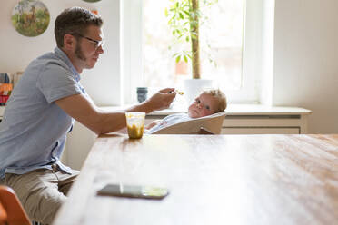 Vater füttert kleinen Jungen, während er zu Hause am Esstisch sitzt - MOEF03104