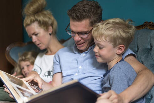 Mann und Sohn sehen sich ein Fotoalbum an, während sie bei einer Frau und ihrer Tochter zu Hause sitzen - MOEF03117