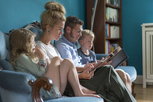 Familie sitzt mit digitalem Tablet und Fotoalbum auf dem Sofa im heimischen Wohnzimmer - MOEF03122
