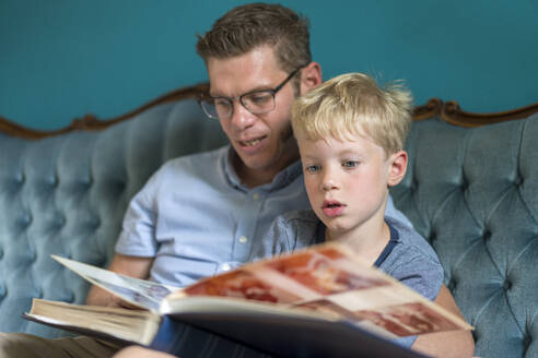 Süßer blonder Junge schaut sich mit seinem Vater ein Fotoalbum an, während er auf dem Sofa im Wohnzimmer sitzt - MOEF03129