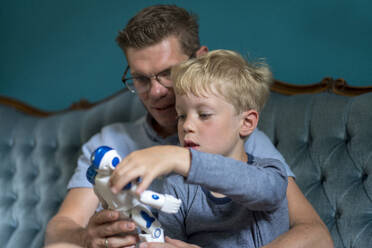 Blond boy looking at robot held by father while sitting on sofa in living room - MOEF03131