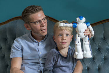 Cute blond boy holding robot while sitting by father on sofa in living room - MOEF03132