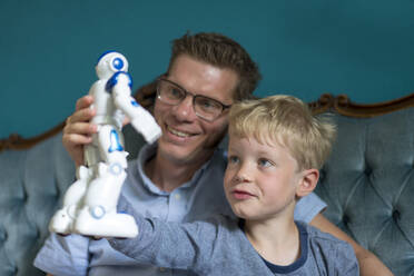 Blond boy looking at robot while sitting by smiling father in living room - MOEF03133