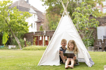 Cute siblings with digital tablet sitting in tent on grass at back yard during weekend - MOEF03140