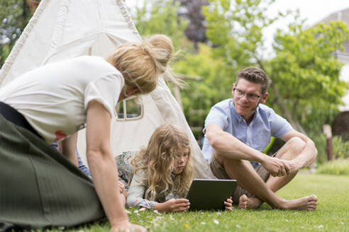 Mädchen benutzt digitales Tablet inmitten der Eltern auf dem Rasen im Hinterhof am Wochenende - MOEF03146