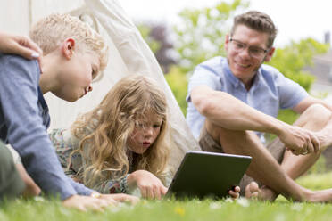 Kids with digital tablet while father sitting on grass at back yard during weekend - MOEF03147