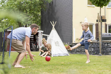 Mann und Sohn spielen Fußball, während die Frau mit ihrer Tochter in einem Zelt im Hinterhof sitzt (Wochenende) - MOEF03148