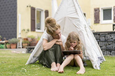 Confused mother and daughter looking at digital tablet while sitting in tent on grass during weekend - MOEF03150