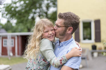 Mann küsst glückliche blonde Tochter, während er sie im Hinterhof trägt - MOEF03162