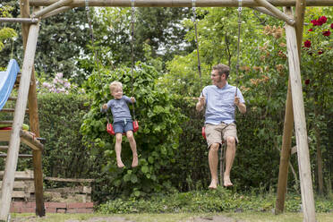 Father and son enjoying swinging against plants at back yard - MOEF03167