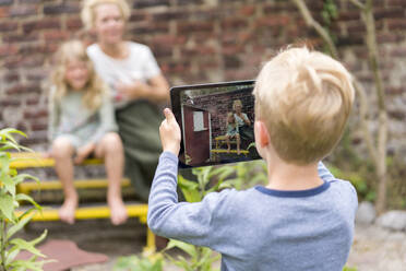 Junge fotografiert Mutter und Schwester durch ein digitales Tablet an einer Backsteinmauer im Hinterhof - MOEF03179