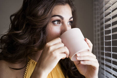 Nahaufnahme einer jungen Frau, die an einer Tasse Kaffee nippt, während sie durch die Jalousien ihres Hauses schaut, lizenzfreies Stockfoto