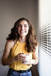 Fröhliche junge Frau, die eine Kaffeetasse in der Hand hält, während sie zu Hause vor den Jalousien steht - EBBF00596