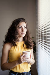 Thoughtful woman holding coffee cup while looking away through blinds at home - EBBF00597
