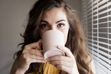 Close-up of young woman with long brown hair drinking coffee by blinds at home - EBBF00598
