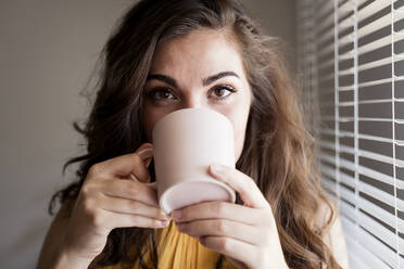 Nahaufnahme einer jungen Frau mit langen braunen Haaren, die zu Hause vor den Jalousien Kaffee trinkt - EBBF00598