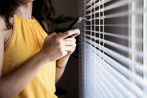 Young woman text messaging on mobile phone by blinds at home - EBBF00601