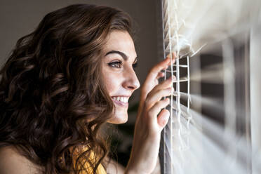 Close-up of happy beautiful young woman looking through window blinds at home - EBBF00603