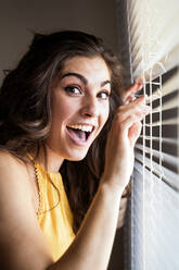 Cheerful young woman with long brown hair standing by blinds at home - EBBF00604