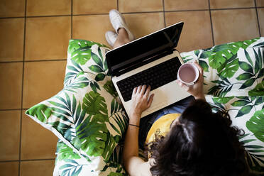 Junge Frau hält eine Kaffeetasse und benutzt einen Laptop, während sie zu Hause auf einem Sessel sitzt - EBBF00605