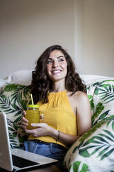 Thoughtful smiling woman holding juice while sitting with laptop on armchair at home - EBBF00610