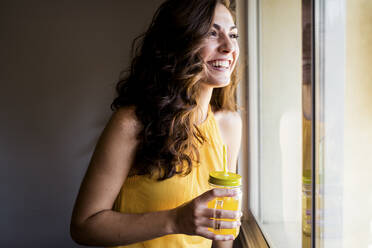 Happy woman holding mason jar with juice while looking away through window at home - EBBF00613