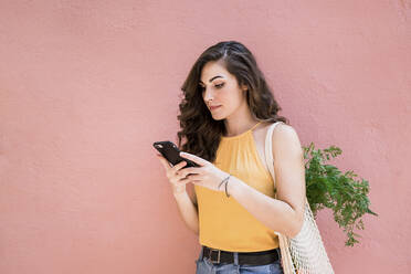 Beautiful young woman using smart phone while standing with reusable mesh bag against pink wall - EBBF00616