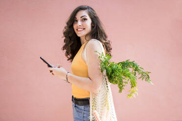 Lächelnde schöne junge Frau, die ihr Smartphone in der Hand hält, während sie mit einer wiederverwendbaren Netztasche vor einer rosa Wand steht - EBBF00618