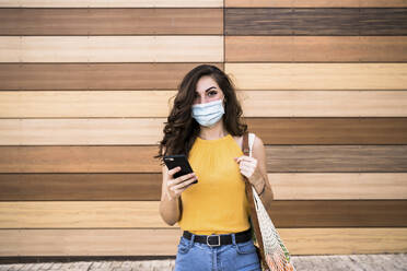 Junge Frau hält Smartphone und Netztasche, während sie an einer Holzwand steht - EBBF00625