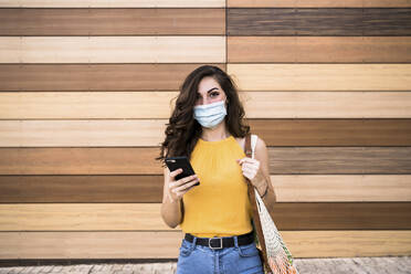 Young woman holding smart phone and mesh bag while standing against wooden wall - EBBF00625