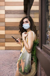 Junge Frau mit Schutzmaske, die ein Mobiltelefon und eine Netztasche hält, während sie an der Wand steht - EBBF00626