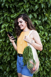 Happy young woman using smart phone while standing with vegetables in mesh bag against green plants - EBBF00635