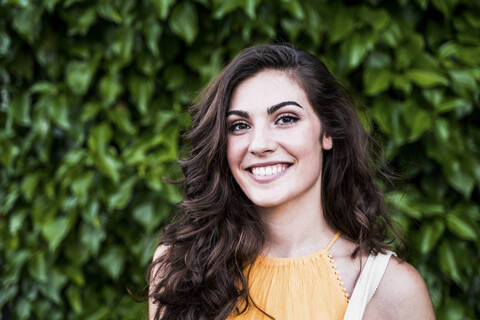 Smiling beautiful young woman with long brown hair standing against plants stock photo