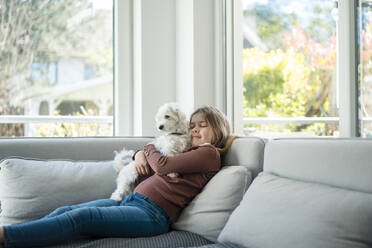 Nettes Mädchen umarmt Hund, während auf dem Sofa im Wohnzimmer sitzen - JOSEF01517