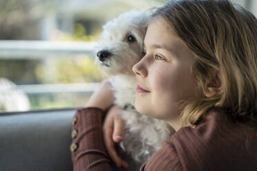 Cute girl embracing dog while looking away in living room - JOSEF01520