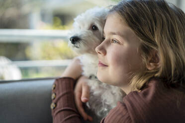 Nettes Mädchen, das seinen Hund umarmt, während es im Wohnzimmer wegschaut - JOSEF01520