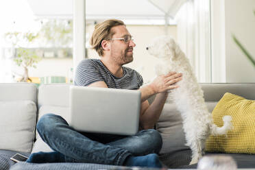 Mann mit Laptop, der einen Hund hält, während er auf dem Sofa sitzt - JOSEF01521