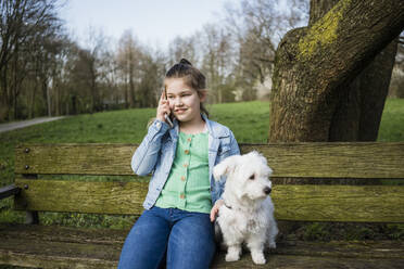 Lächelndes Mädchen mit Hund telefoniert auf einer Bank im Park - JOSEF01538