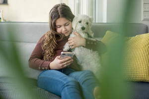Mädchen benutzt Smartphone und hält Hund auf Sofa - JOSEF01560