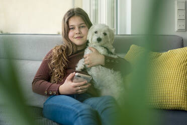 Smiling girl using smart phone while holding dog on sofa - JOSEF01561