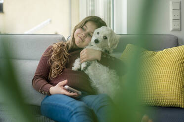 Mädchen mit Smartphone schaut weg und hält Hund auf Sofa - JOSEF01562