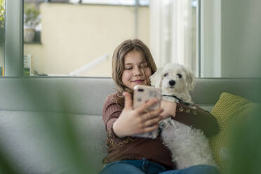 Mädchen nimmt Selfie durch Smartphone mit Hund auf Sofa - JOSEF01563