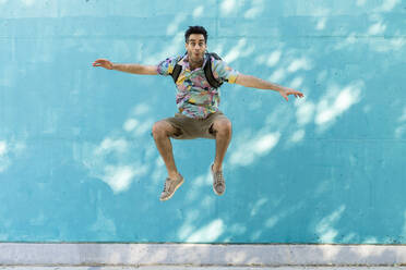 Happy man with backpack jumping in front of blue wall - AFVF06967